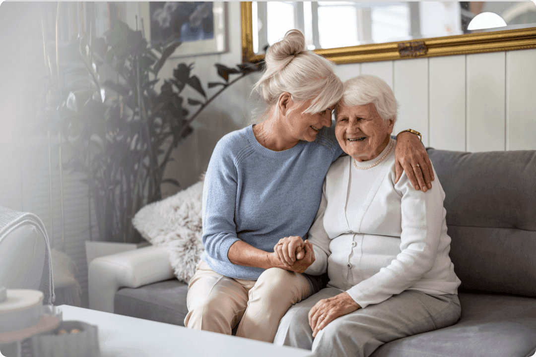 Two elderly women sitting on a couch share a joyful moment, with one woman affectionately holding the other's hand.